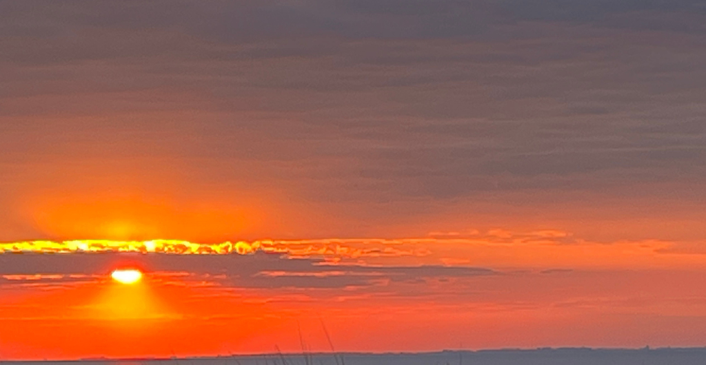 Vor grauviolettem Himmel im oberen Drittel geht die Sonne leuchtend hinter einem orangeroten Wolkenband unter. von K. Schwahlen lizensiert unter CC BY-SA